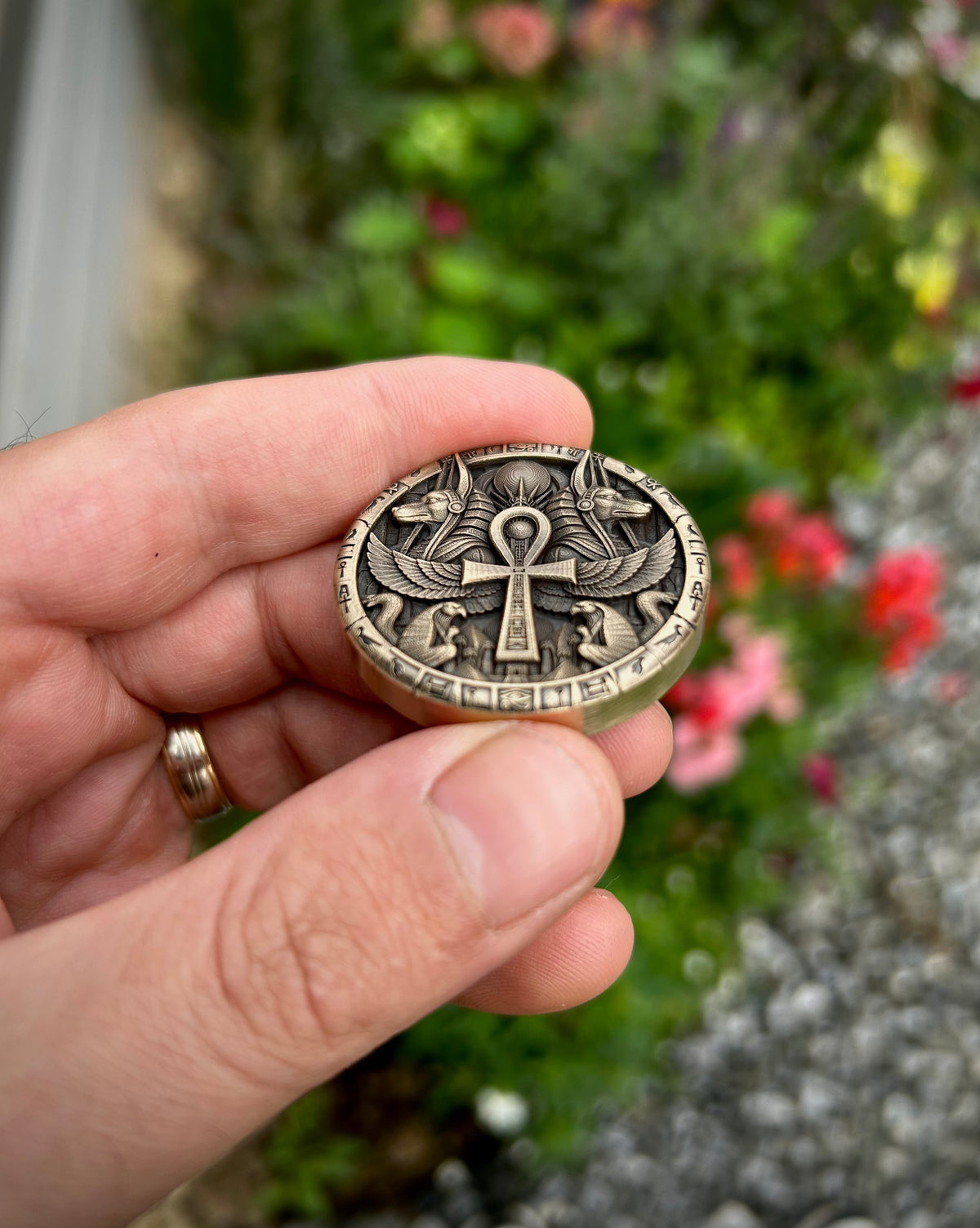 A hand holding a round antique-style metal medallion featuring an Egyptian ankh key design, with a blurred garden background.