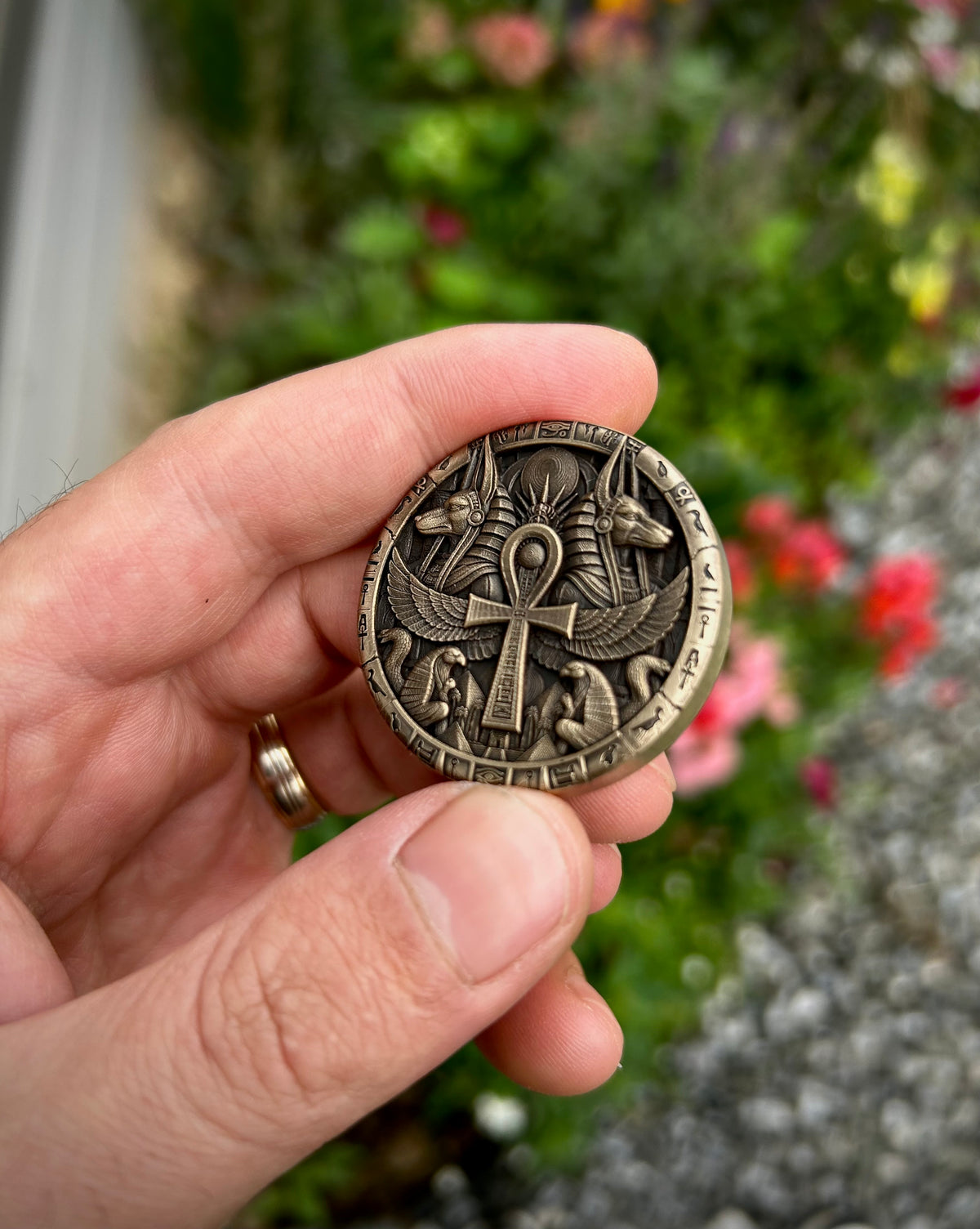 A hand holding a round bronze-toned coin featuring Egyptian motifs, including an ankh and winged scarab beetle, with a blurred garden background.