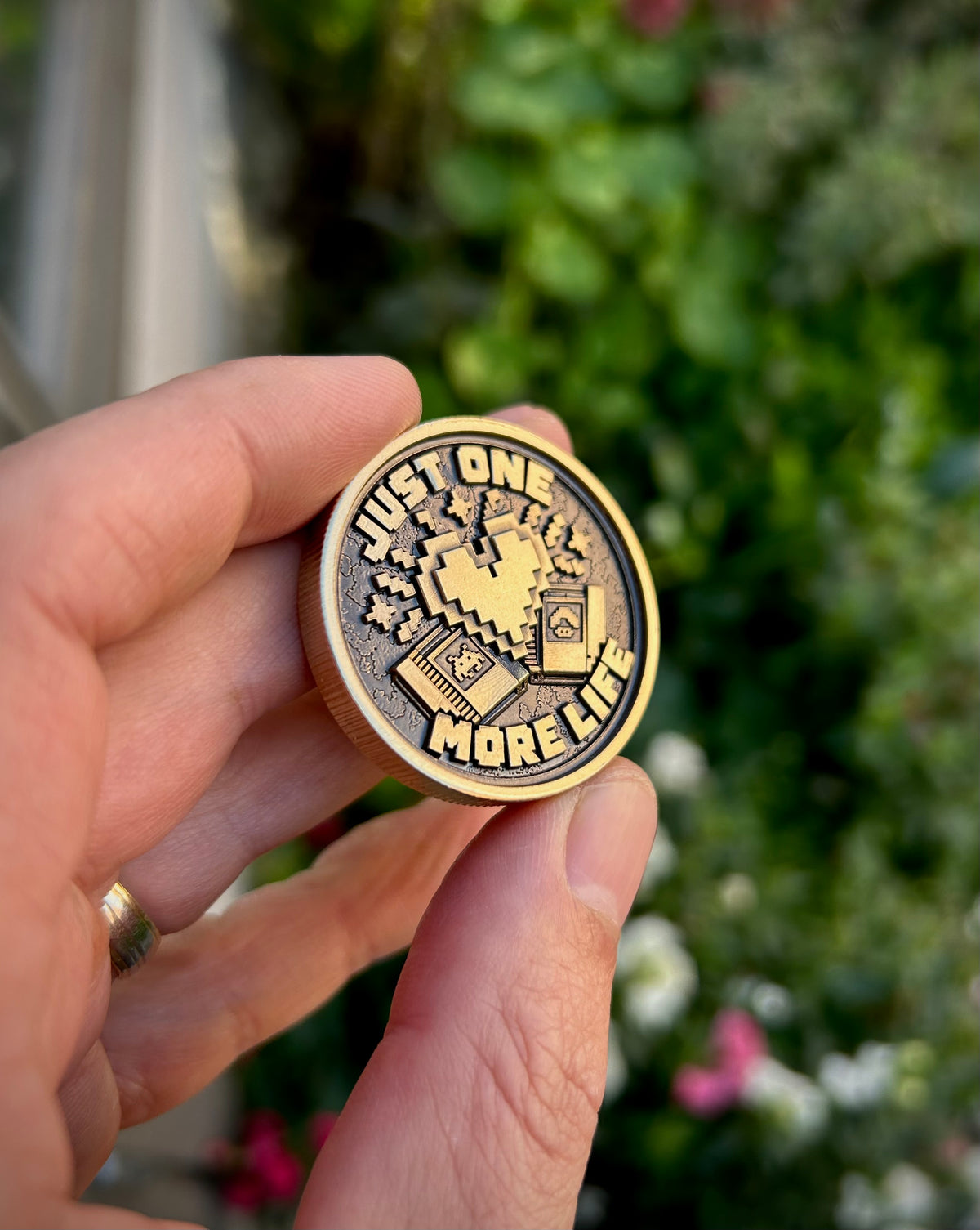 Just One More Life pocket coin held at an angle, showing its pixel heart, two retro cartridges, raised lettering and ridged edge.