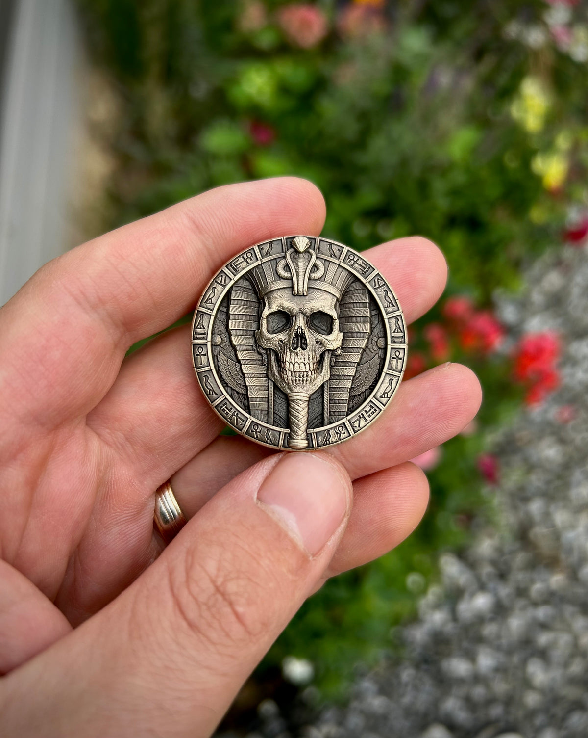 A hand holding a round antique bronze coin featuring a detailed Egyptian skull and scarab design, with a blurred garden background.