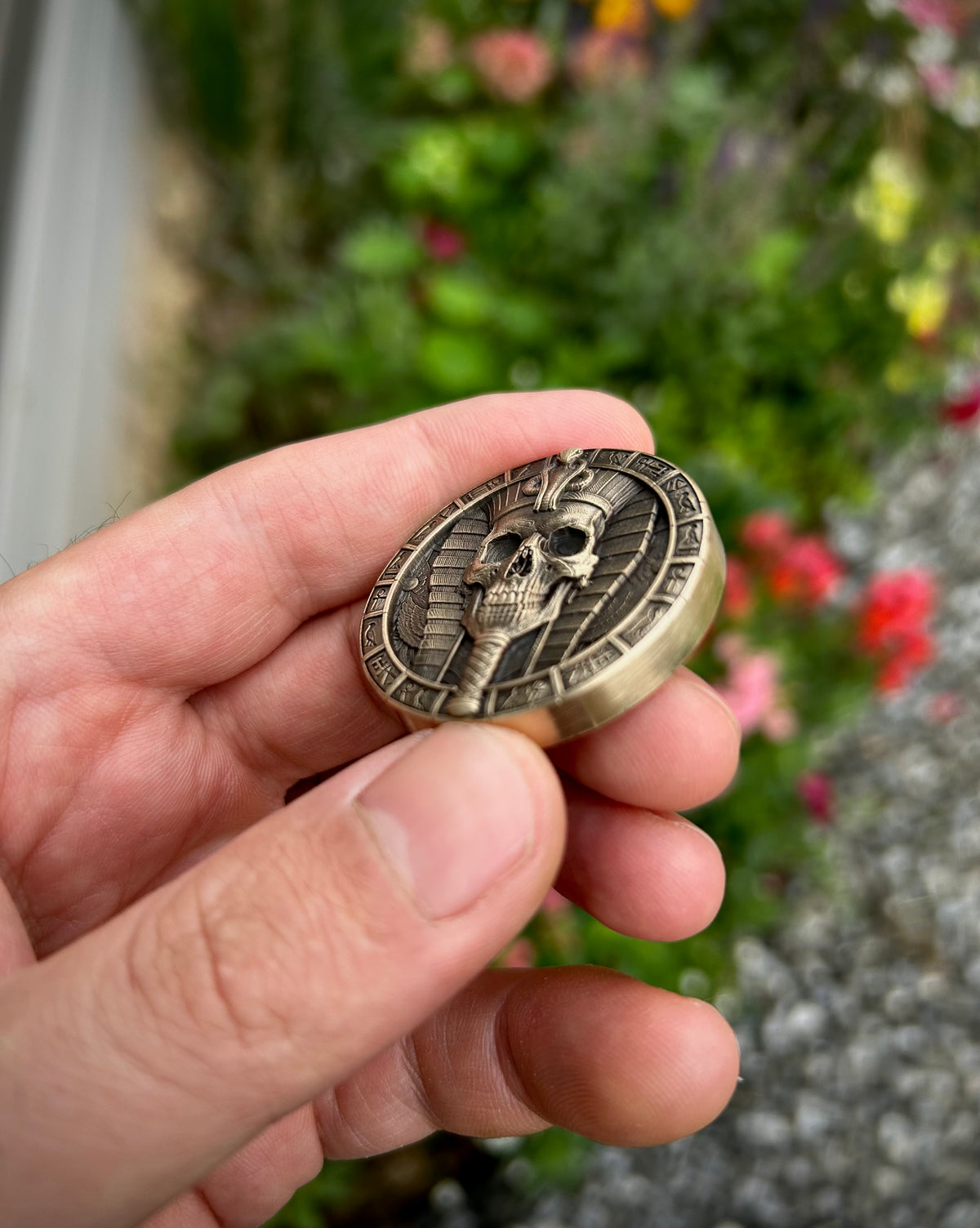 A hand holding a round bronze-toned metal medallion featuring a skull and cartouche design, with a blurred garden background.