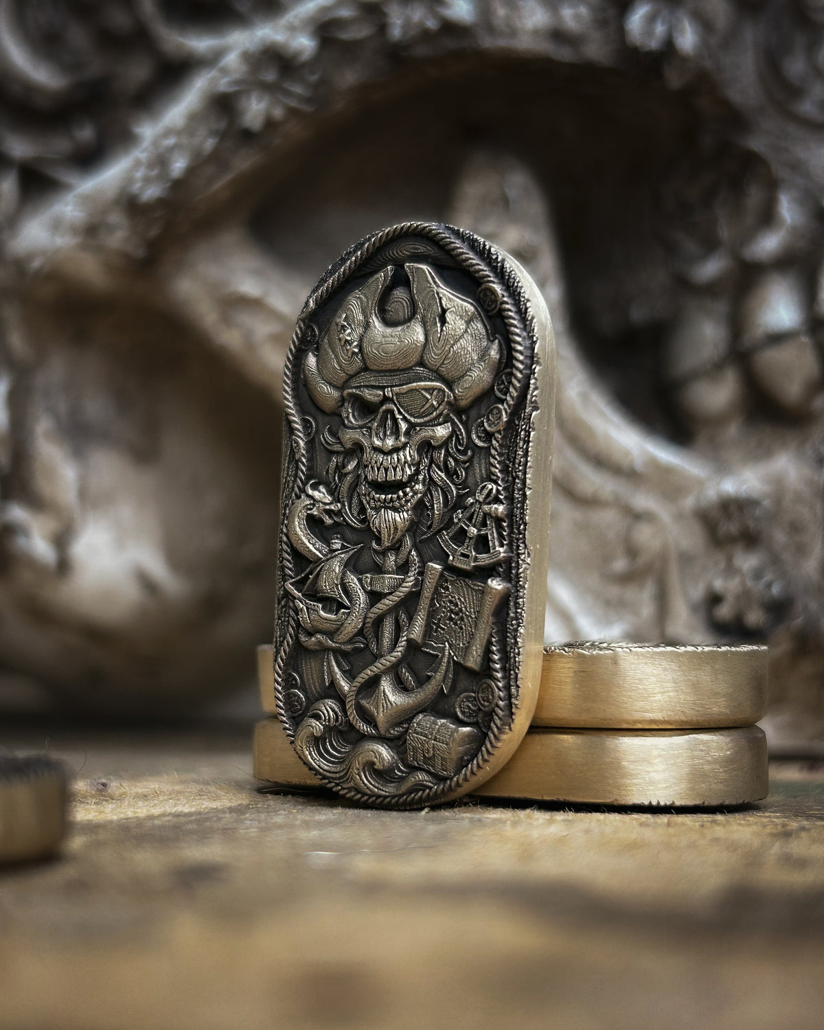 An antique bronze pirate skull pendant or ring holder displayed upright on stacked gold-toned rings, with a blurred skull prop in the background.