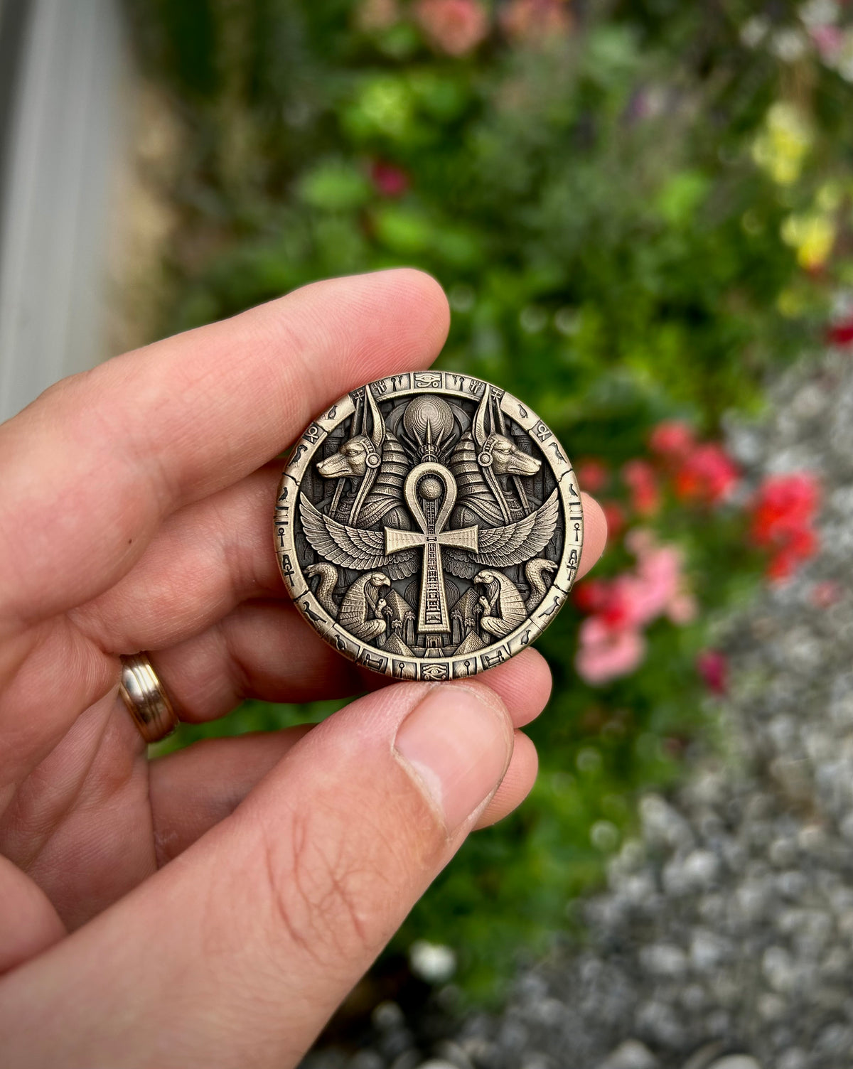A hand holding a round metallic coin or medallion featuring an Egyptian ankh cross design, with a blurred garden background.