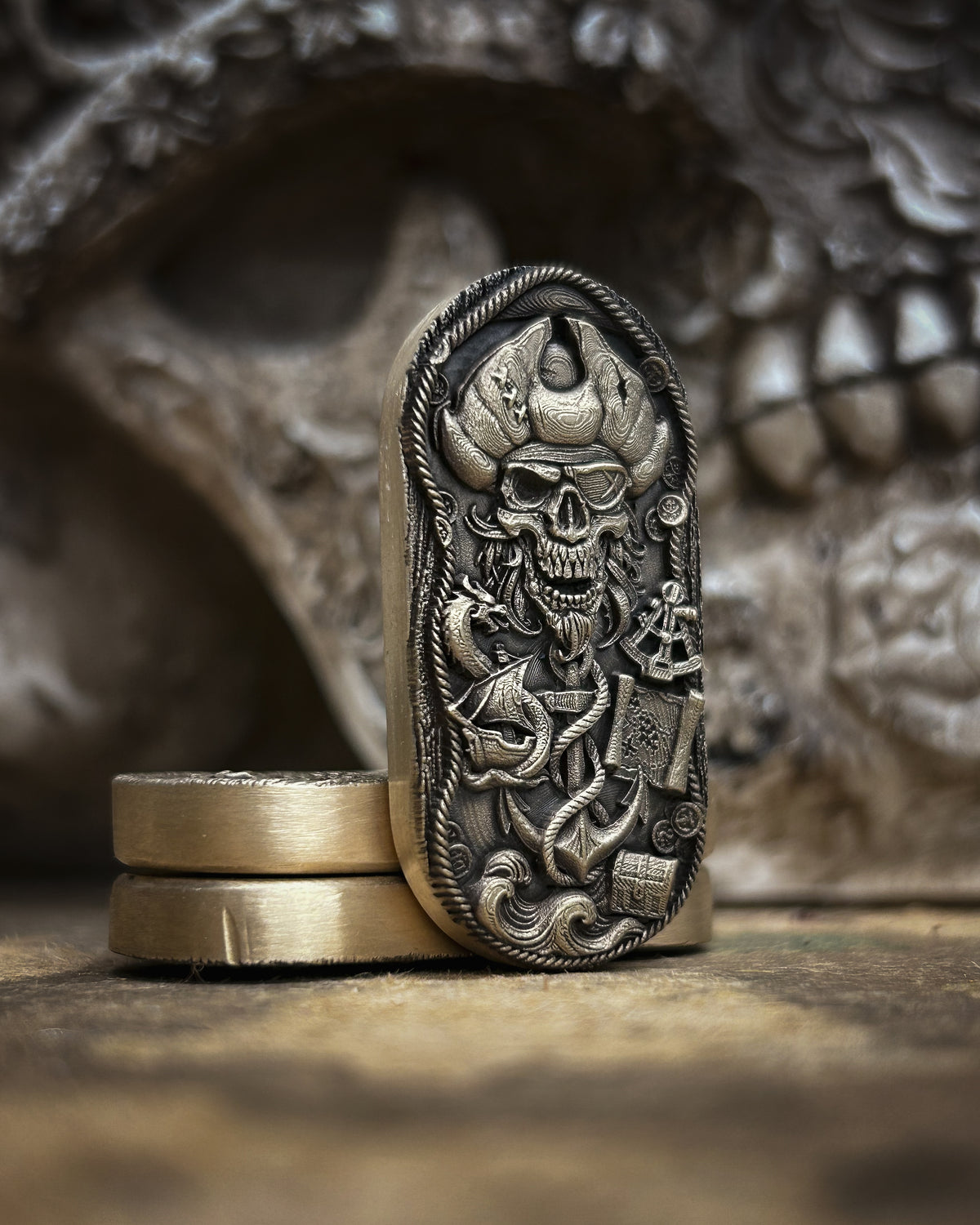 An ornate silver-toned metal pendant or bookmark featuring a detailed skull and pirate-themed engraving, standing upright on two gold-toned rings against a dark, blurred skull prop background.