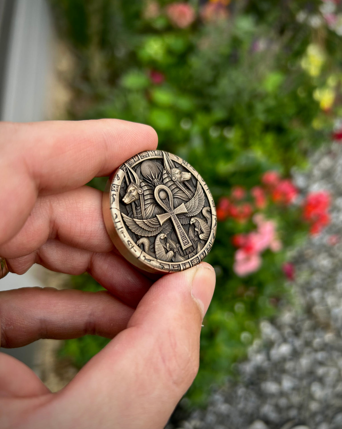 A hand holding a small round bronze-toned metal charm or medallion with an engraved ankh cross and bird design, set against a blurred garden background.