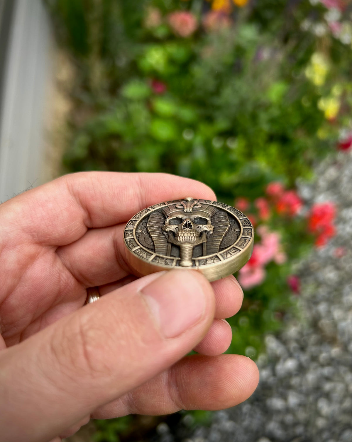 A hand holding a round antique bronze coin featuring a detailed skull and cartouche design, with a blurred garden background.