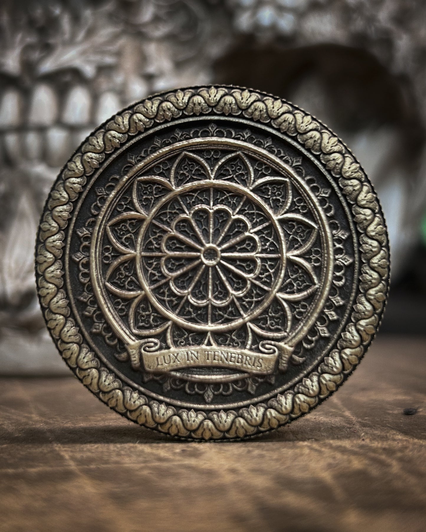 Decorative metal coin with intricate design and 'Lux in Tenebris' text on a wooden surface.