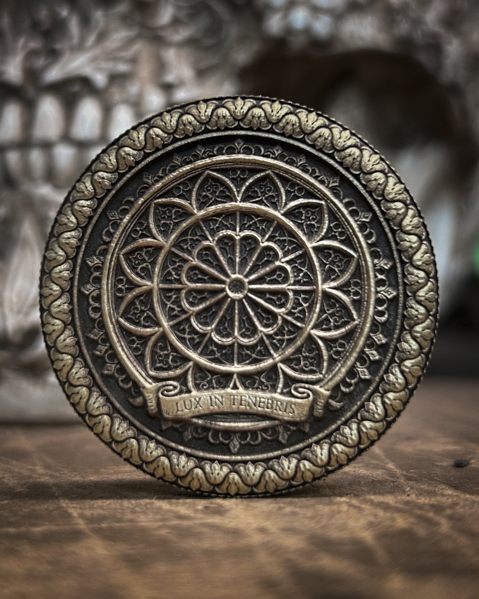Decorative metal coin with intricate design and 'Lux in Tenebris' text on a wooden surface.