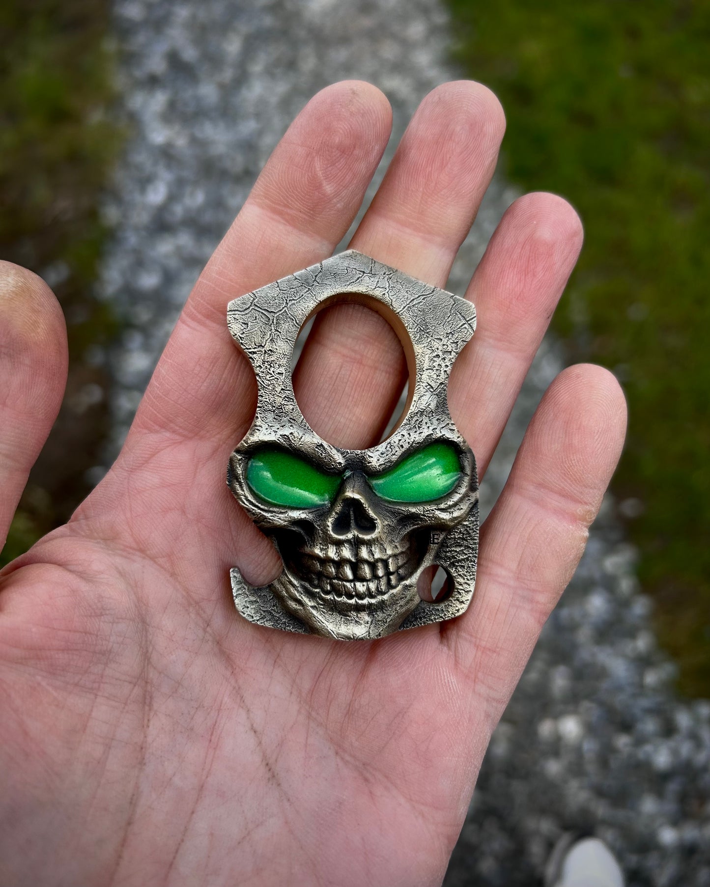 Skull-shaped bottle opener with green glow in the dark eyes held in a hand against a natural background