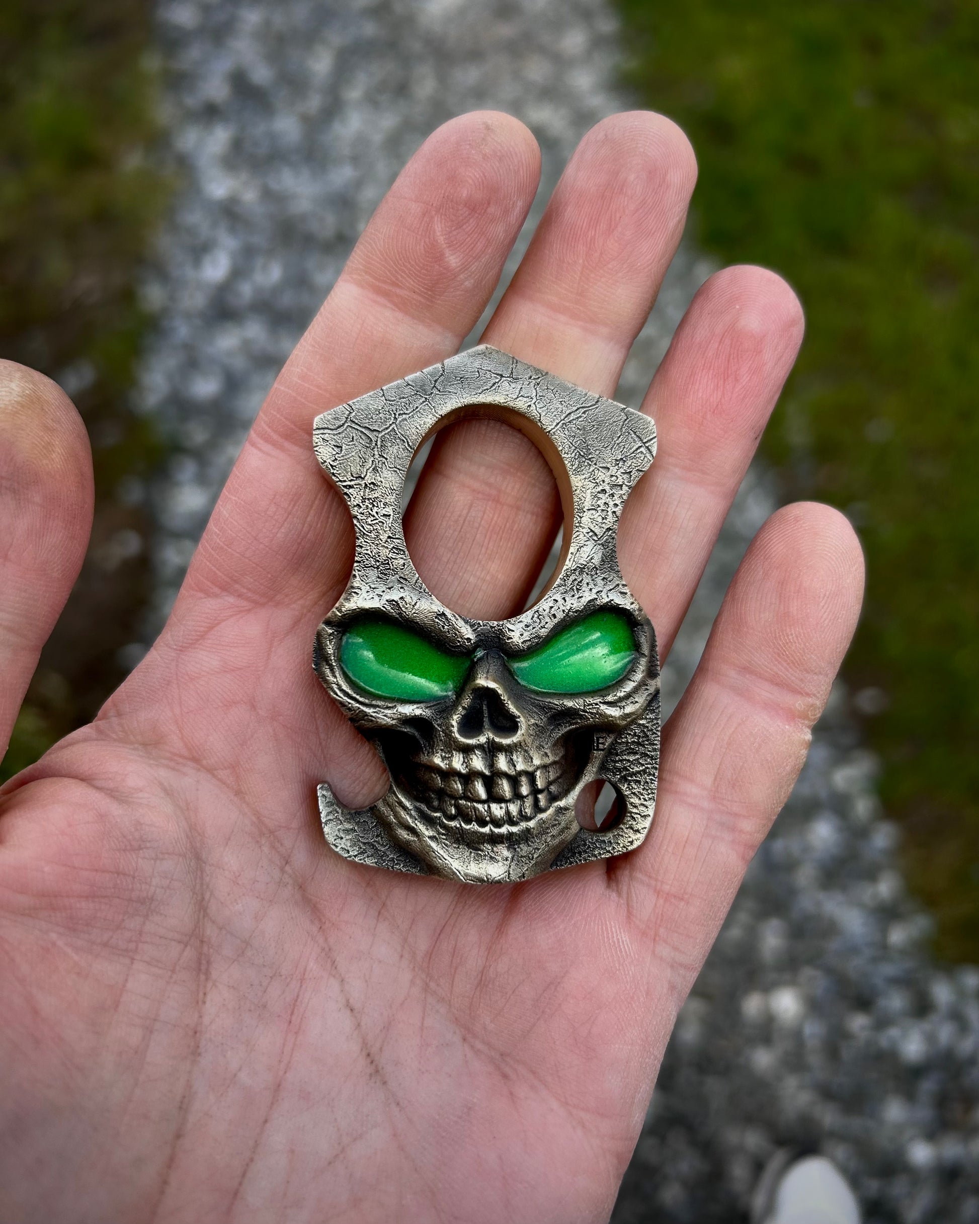Skull-shaped bottle opener with green glow in the dark eyes held in a hand against a natural background
