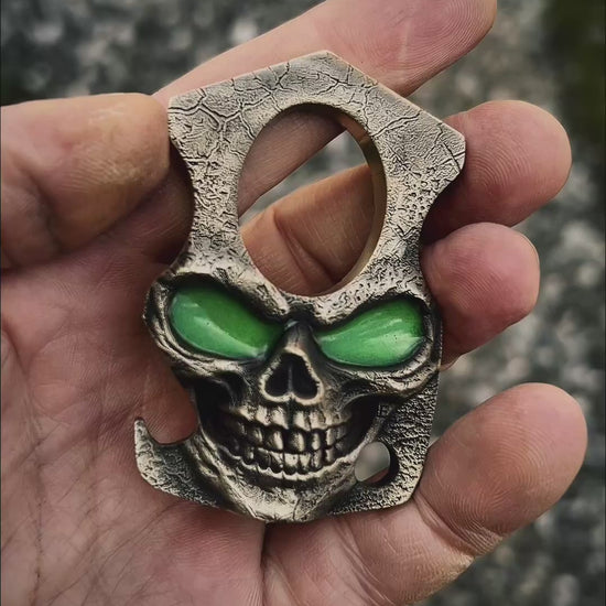 Skull-shaped bottle opener with green glow in the dark eyes held in a hand against a natural background