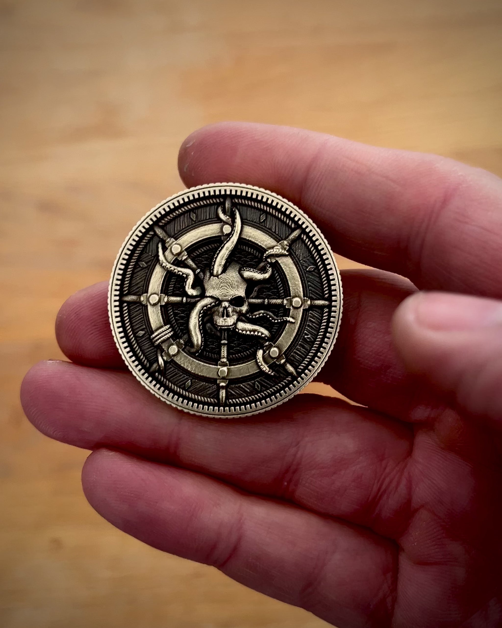Hand holding a brass coin with a detailed pirate themed design against a wooden background